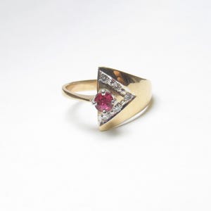 Op de afbeelding: Een gouden ring met een driehoekig ontwerp. De ring heeft een centrale, ronde, rode edelsteen, omringd door kleinere, heldere stenen. De band van de ring is van eenvoudig, gepolijst goud.
