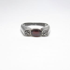 May include: A silver ring with a central oval, dark red gemstone and two smaller, circular decorative elements on either side. The ring has a simple, elegant design, suitable for everyday wear.