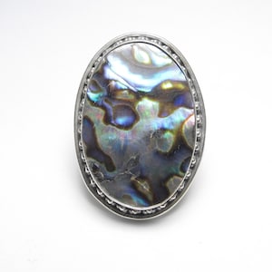 May include: An oval ring featuring a large, iridescent abalone shell inlay with swirling patterns of blue, green, and purple. The shell is set within a polished silver-toned metal frame that is decorated with a beaded texture along the outer edge.