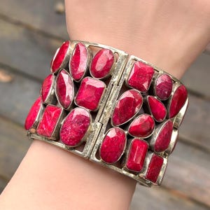 May include: A silver bracelet featuring multiple vibrant red gemstones in various shapes. The gemstones are set in a rectangular silver frame, creating a bold and eye-catching design. The bracelet is a statement piece of jewelry.