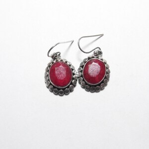 May include: A pair of silver earrings with oval, faceted, ruby-red gemstones. Each earring has a silver border with small, round accents. The earrings have a simple hook design for pierced ears.