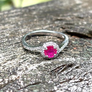 May include: A silver-toned ring featuring a round, vibrant pink center stone surrounded by a halo of small, clear, sparkling stones. The band is slender and also encrusted with a row of tiny, clear, reflective stones along the visible shank.