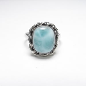 May include: A silver ring featuring a large, oval-shaped, light blue cabochon stone with white marbled patterns. The stone is set within a decorative, twisted rope-style metal border, and the band is a smooth, polished silver metal finish.