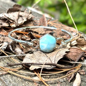May include: A silver bangle bracelet featuring a hexagonal, light blue gemstone. The gemstone is the focal point, connected to the bracelet with small silver loops. The bracelet rests on a bed of dried leaves and wood.