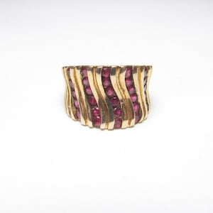 Op de afbeelding: Een goudkleurige ring met een golvend ontwerp. De ring is gevuld met rijen kleine, ronde, rode edelstenen. Het ontwerp van de ring heeft verticale, gebogen gouden banden die een textuur creëren.