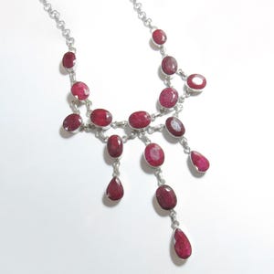 May include: A silver necklace featuring multiple faceted, deep red oval and teardrop-shaped ruby gemstones set in polished metal bezels. The design includes a central drop arrangement with interconnected links, creating a cascading, layered appearance.