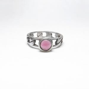 May include: A silver ring with a chain-link design. The ring features a round, pink gemstone set in the center. The ring is displayed on a white background.