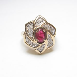 May include: A gold ring featuring a central oval-cut red ruby held by four prongs. The ruby is surrounded by a swirling, star-shaped frame set with numerous small, clear, round-cut diamonds, creating a layered, floral-inspired design.