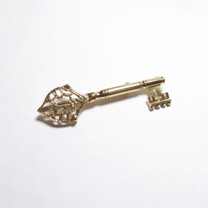 May include: A gold-colored decorative key with an ornate, openwork design on the head. The key has a detailed, vintage aesthetic, with a textured handle and a classic key bit. The key is set against a white background.