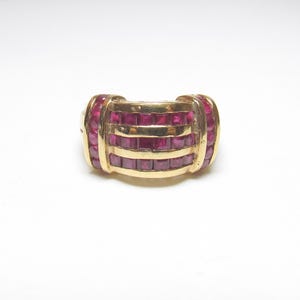 May include: A gold ring with a rectangular design, featuring multiple rows of square-cut rubies. The ring has a rounded shape with the gemstones set in a channel style. The gold band is visible between the rows of rubies.