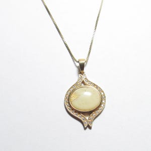 May include: Gold-toned pendant necklace featuring an oval, cream-colored stone surrounded by small, clear crystals. The pendant has a decorative, openwork design and hangs from a delicate chain. A classic piece of jewelry.