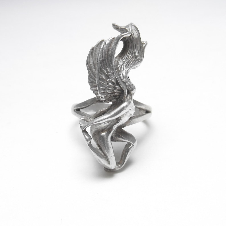 Puede incluir: Un anillo de plata con una figura alada esculpida. El anillo presenta un dise&ntilde;o detallado de una persona con alas, creando una joya &uacute;nica y art&iacute;stica. El anillo est&aacute; sobre un fondo blanco, resaltando los detalles.