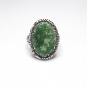 May include: Oval-shaped ring featuring a large, green stone with white marbling. The stone is set in a silver-toned metal with a twisted rope-like border. The ring has a simple band.
