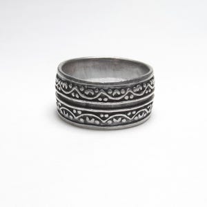 May include: A wide silver-toned metal ring featuring a dark, oxidized finish. The band is decorated with two parallel horizontal rows of embossed, repeating wave-like patterns interspersed with small raised dots along the entire circumference.
