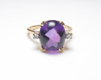 May include: A gold ring featuring a large, oval-cut, deep purple amethyst gemstone held by four prongs. The band is accented on each side by a single, small, round-cut clear diamond set in a gold mounting, creating a balanced and elegant design.