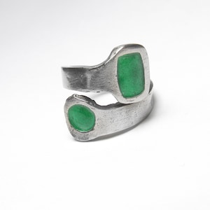 May include: A silver-toned ring with two sections, each featuring a bright green, rectangular or oval-shaped stone. The ring has a textured, slightly worn appearance, with the stones set into the metal.
