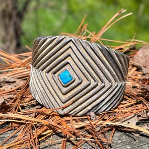 May include: A silver-toned cuff bracelet with a geometric design featuring a central square with a turquoise stone. The bracelet has a textured surface and is set against a natural background of pine needles.