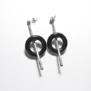 May include: A pair of modern earrings featuring a black circular design with a silver bar and post. The earrings have a post back and a small silver ball at the top. The earrings are set against a white background.