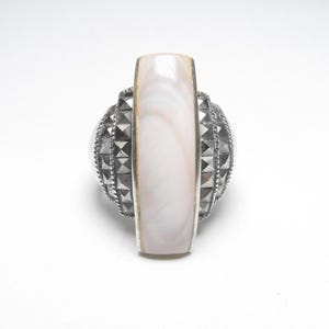 May include: A ring featuring a long, rectangular, iridescent pale pink mother-of-pearl inlay set vertically. The band is crafted from dark, metallic, faceted stones arranged in a pyramid-like texture, creating a geometric and layered appearance.