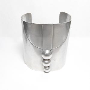 May include: A wide, silver cuff bracelet. The bracelet has a curved top and a vertical rectangular accent in the centre. Below the accent are three graduated, spherical embellishments. The bracelet is made of a shiny metal.