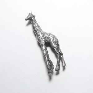 May include: A silver-colored giraffe figurine with a textured surface. The giraffe has a long neck, small ears, and detailed hooves. The body is covered in a pattern of raised circles. The figurine is set against a white background.
