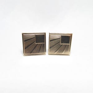 May include: A pair of gold-toned cufflinks. Each cufflink features a square design with a smaller square in the upper corner and radiating lines. The cufflinks are set against a white background.