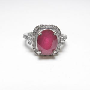 Op de afbeelding: Een zilveren ring met een grote, ovaal geslepen robijn. De robijn heeft een dieprode kleur en is omgeven door een halo van kleinere, heldere edelstenen. De ring heeft decoratieve filigraan details op de band.