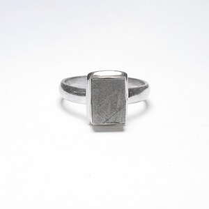 May include: A silver ring with a rectangular, textured face. The ring band is smooth and rounded, leading to the prominent rectangular centerpiece. The ring is set against a white background, highlighting its metallic sheen.