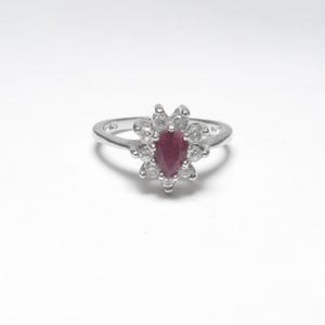 May include: A silver ring featuring a central, pear-shaped, deep red gemstone, surrounded by a halo of smaller, clear gemstones. The ring's band is thin and polished, with a simple design. The ring is set against a white background.