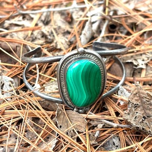 May include: A silver cuff bracelet with a large, oval malachite stone. The stone is a vibrant green with swirling patterns. The bracelet is set against a backdrop of dried pine needles and leaves.