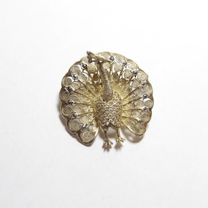 May include: A gold peacock brooch with intricate filigree details. The peacock's tail feathers are spread out in a fan shape.