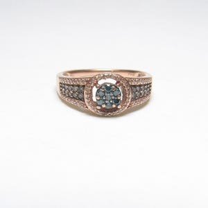 Op de afbeelding: Roségouden ring met een centrale cluster van blauwgroene stenen, omringd door een halo van kleinere stenen. De band is versierd met extra stenen, wat een sprankelend effect creëert. Een klassiek en elegant sieraad.