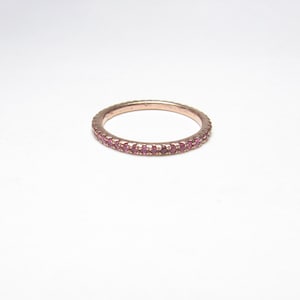 May include: A rose gold ring featuring a band of small, round, pink gemstones. The ring is set against a white backdrop, highlighting the intricate details of the jewellery. The gemstones are evenly spaced around the band.