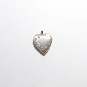 May include: A heart-shaped silver locket pendant featuring a matte finish and a series of small, engraved leaf-like patterns arranged in a curved line along the upper portion of the front surface, complete with a small metal bail at the top.