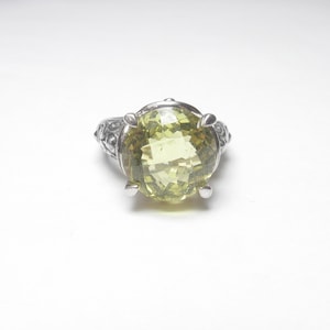 May include: A silver ring featuring a large, round, pale green gemstone set in a claw setting. The band of the ring is adorned with intricate details.