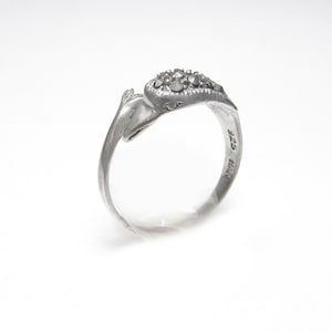 May include: A silver ring with a heart-shaped design, adorned with small, dark gemstones. The ring has a smooth, polished band and a decorative element on the side. The ring is set against a white background.