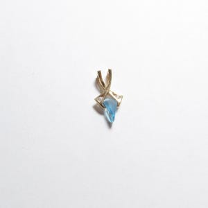 May include: A gold pendant featuring a triangular, translucent light blue stone set within a polished gold frame that crosses at the top. Two small, clear, round-cut stones are embedded in the gold setting on either side of the blue stone.