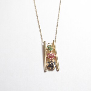 May include: Gold-toned necklace featuring a ladder-like pendant with three colorful gemstone clusters. The top cluster is green, the middle is pink, and the bottom is dark blue. The necklace has a delicate chain.