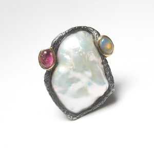 May include: A statement ring with a large, irregular white pearl in a textured silver setting. It features a pink gemstone and an opal, each in gold bezels. The ring's design is modern and artistic.