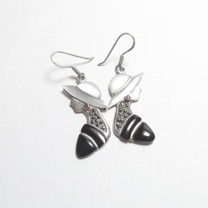 May include: A pair of silver earrings featuring a black and white cameo design. Each earring has a woman's profile with a hat, a black onyx stone, and a small red gemstone.