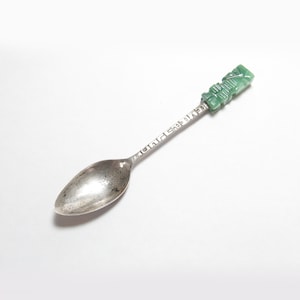 May include: A silver spoon with a green jade handle. The handle is carved with a design that resembles a tiki figure. The spoon is engraved with the words "Island Shop Oahu".