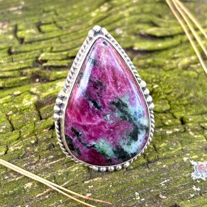 May include: A teardrop-shaped ring featuring a polished ruby zoisite gemstone with vibrant magenta and dark green inclusions. The stone is set in a sterling silver bezel with a decorative twisted rope border and small silver bead accents.