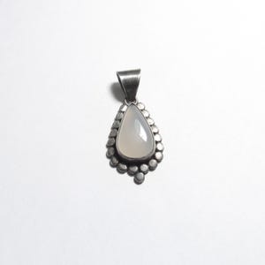 May include: A silver pendant with a pear-shaped, pale-colored stone. The stone is surrounded by a beaded silver border and has a silver loop at the top for a chain. The pendant is set against a white background.