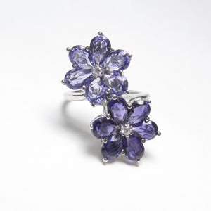 May include: A silver ring featuring two flower-shaped clusters of faceted gemstones. The top flower consists of light purple pear-shaped stones with a small clear center stone, while the bottom flower has dark purple pear-shaped stones.
