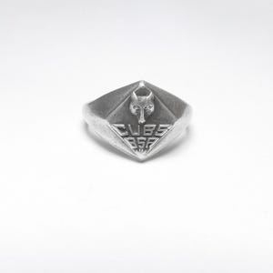May include: Silver-toned ring with a diamond-shaped face. The face features a wolf's head above the text "CUBS BSF". The ring is likely made of metal and has a vintage or antique appearance. The design suggests a fraternal or scouting theme.
