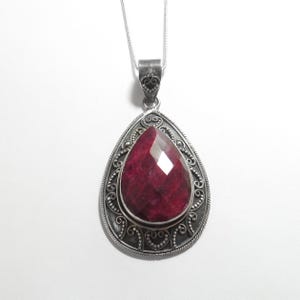 May include: A silver pendant necklace with a large, faceted, teardrop-shaped ruby gemstone. The ruby is set in an ornate, filigree-style silver frame. The pendant hangs from a delicate silver chain, creating an elegant piece of jewellery.