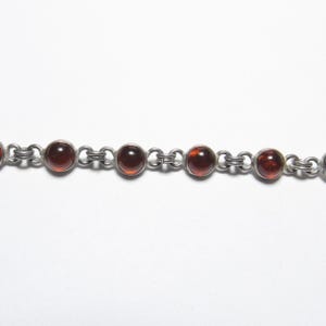 May include: A silver chain bracelet featuring round, reddish-brown gemstones. The gemstones are evenly spaced and connected by small, linked silver chain segments. The bracelet is set against a plain white background.