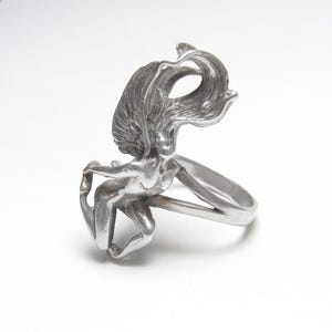 Puede incluir: Un anillo plateado con una figura femenina esculpida con cabello ondeante y brazos extendidos. El dise&ntilde;o del anillo integra la forma de la mujer en la banda. El anillo est&aacute; hecho de un material met&aacute;lico brillante.