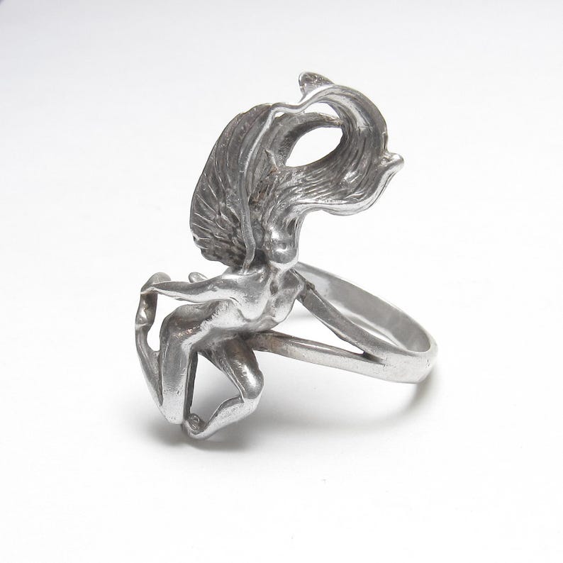 Puede incluir: Un anillo plateado con una figura femenina esculpida con cabello ondeante y brazos extendidos. El dise&ntilde;o del anillo integra la forma de la mujer en la banda. El anillo est&aacute; hecho de un material met&aacute;lico brillante.