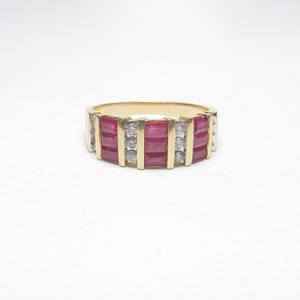 May include: A gold ring featuring a band of rectangular, deep red gemstones and small, clear stones. The ring has a classic design, with the red stones set between vertical gold bars. The ring is set against a white background.